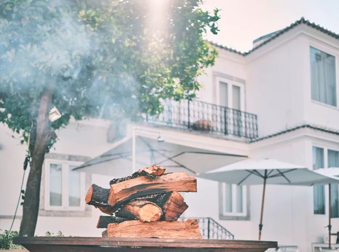 A stack of wood resting on a grill, set against the backdrop of a building's facade and a large tree.