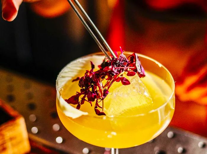 A hand delicately places a colorful array of edible flowers atop a vibrant cocktail in a coupe glass using tweezers.