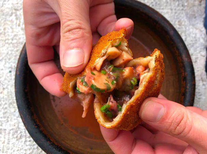 Hands hold a croquette, split open to reveal savory meat and vegetables inside, presented on a dark ceramic plate.