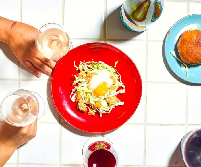 An overhead view of a tiled table featuring a plate of cabbage salad topped with croutons and a runny egg, alongside a sandwich and hands holding glasses of red and white wine.