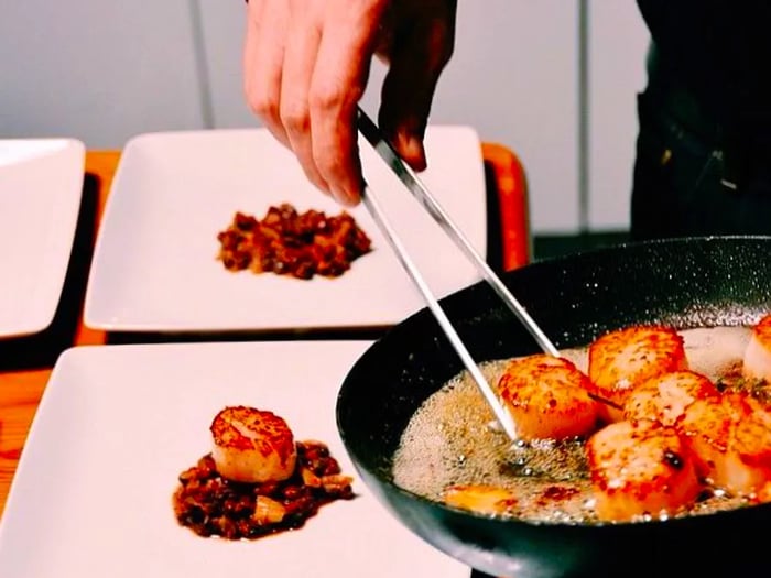 A hand carefully uses tongs to move cooked scallops from a pan to minimalist plates.