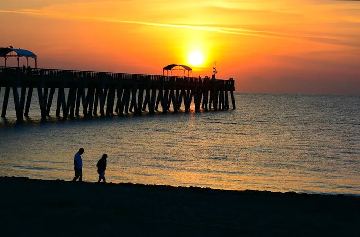 A sunrise illuminating a beach pier with two adults silhouetted walking along the shore