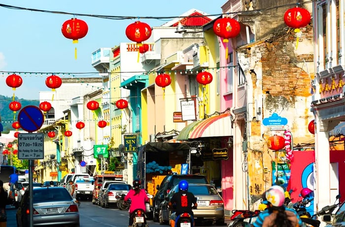 Motorcyclists ride along a narrow street lined with historic shophouses, adorned with hanging lanterns overhead.