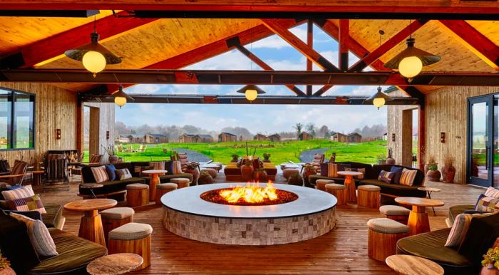 A spacious communal area featuring light wood sofas, tables, and stools arranged around a tile-lined fire pit, complemented by stylish pendant lights suspended from a tall ceiling with exposed rafters. The far wall opens up to the surrounding landscape.