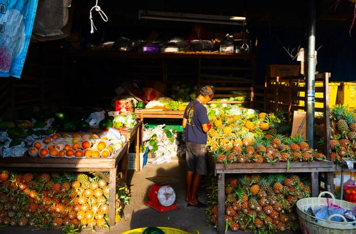 A vendor stands amidst a blend of sunlight and shade, surrounded by tables laden with pineapples and citrus fruits.