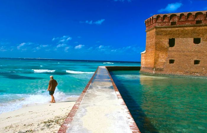 A lone figure wades into the sea on a beach beside a historic fortress.