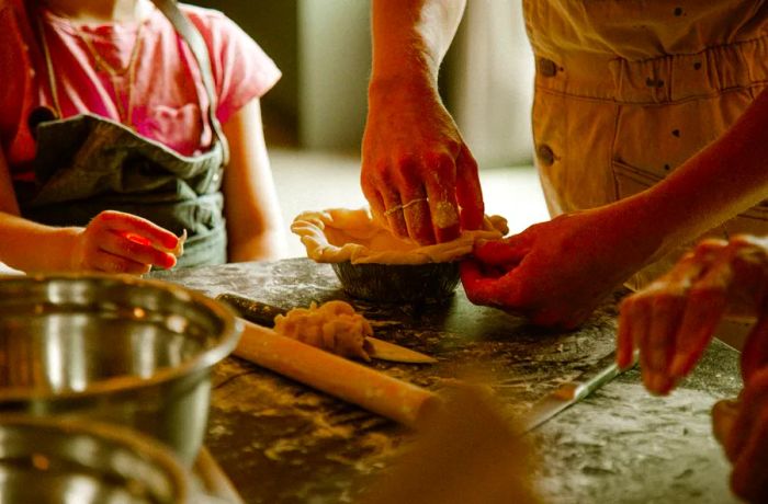 An adult shapes the edges of a pie crust in a tin, while a child observes nearby.