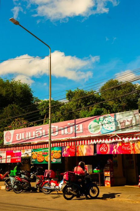 Motorcyclists are parked outside a market that promotes Thai food under a clear blue sky.