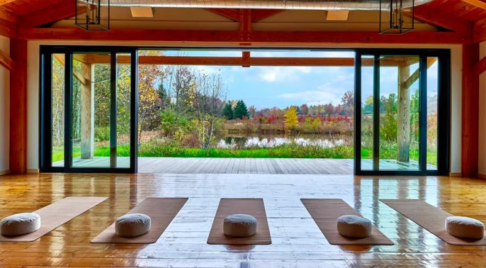 Yoga mats are arranged on a spacious wooden floor inside a tall wooden structure, which features a large wall opening to the outdoors.