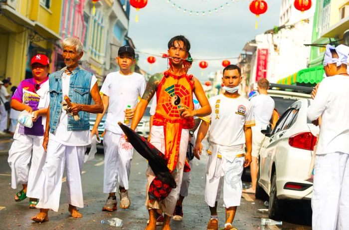 A group of individuals strolls down the street, most dressed in white attire. A leader among them has long skewers pierced through their cheeks.