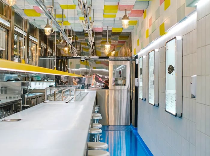 A spacious dining area featuring a long counter, blue flooring, tiled walls, vibrant ceiling tiles, and an abundance of shiny silver metal surfaces.