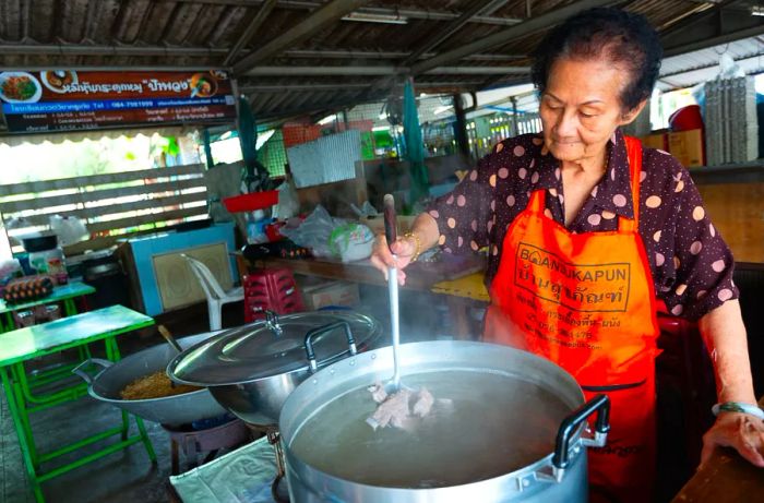 An elderly chef stirs a large pot over the heat.