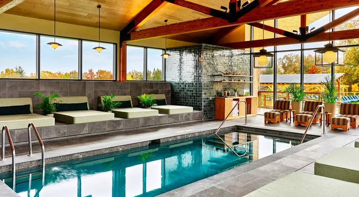 A cozy pool area encircled by large daybeds made of concrete and additional seating, all beneath a lofty ceiling with exposed beams.