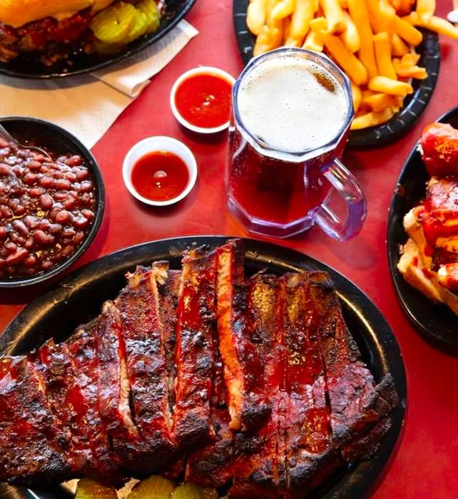 A table laid out with a large platter of ribs, a small bowl of baked beans, a mug of beer, fries, a burnt ends hoagie, and various sauces.