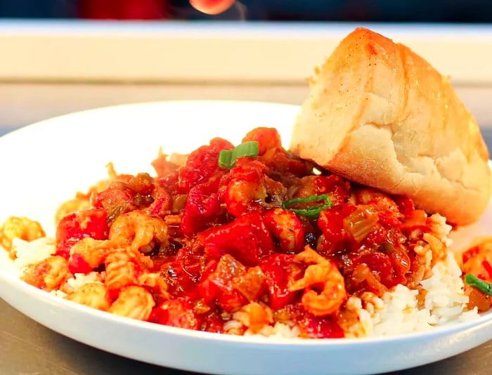 A chef meticulously adds a garnish to a bowl of crawfish etouffee, accompanied by a generous piece of bread.