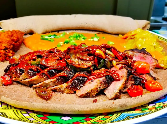 Slices of steak topped with roasted vegetables and awaze sauce, served on a bed of injera alongside an array of sides.