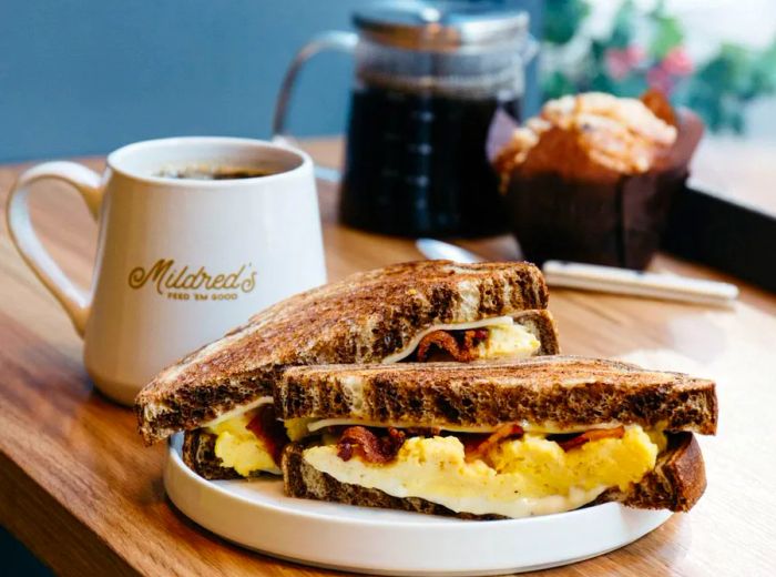 A sandwich cut in half, overflowing with eggs and bacon, served on dark rye bread alongside a branded coffee mug.