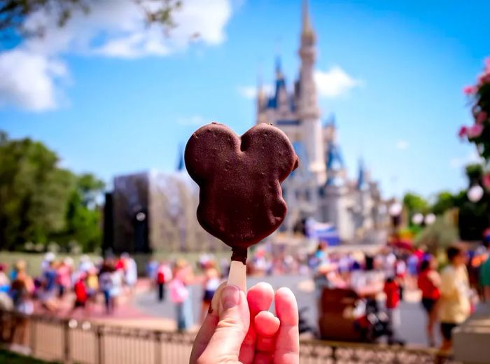 A hand holds a chocolate treat crafted to resemble Mickey Mouse's head in front of the Magic Castle.