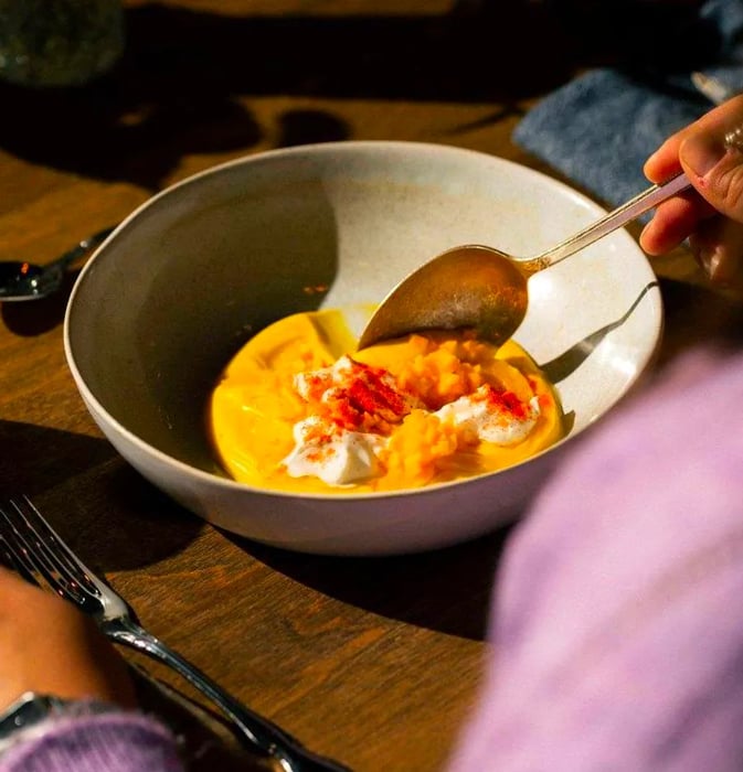 A diner scoops a spoon into a bowl filled with thick yellow sauce, topped with creamy cheese and other delicious toppings.