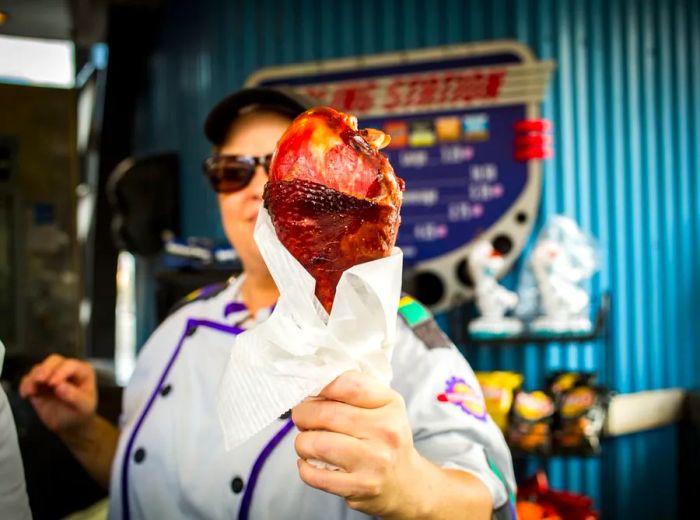 A server offers a large turkey leg, wrapped in wax paper, towards the camera, with a menu and snacks blurred in the background.