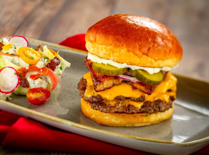 A towering cheeseburger bursting with cheese, pickles, onions, and sauce, accompanied by a small salad on a metal tray.