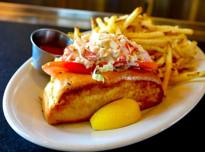 A generously filled lobster roll served with french fries, accompanied by a lemon wedge and a ketchup container on the plate.