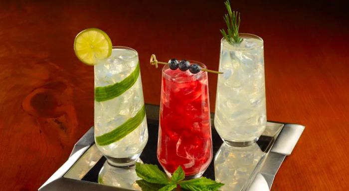 Three highball cocktails displayed on a silver tray: one garnished with cucumber and lemon, another a vibrant red with blueberries, and a fizzy one topped with thyme.