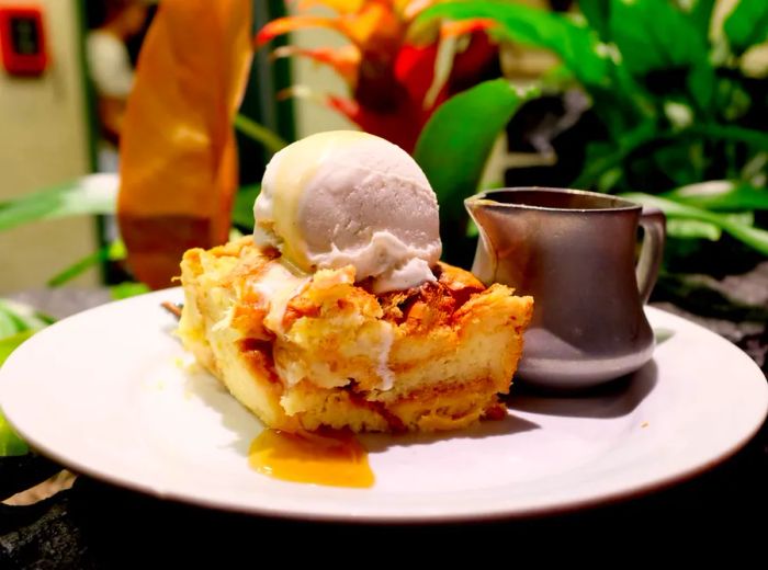 A generous piece of bread pudding, adorned with ice cream and accompanied by a carafe of syrup.