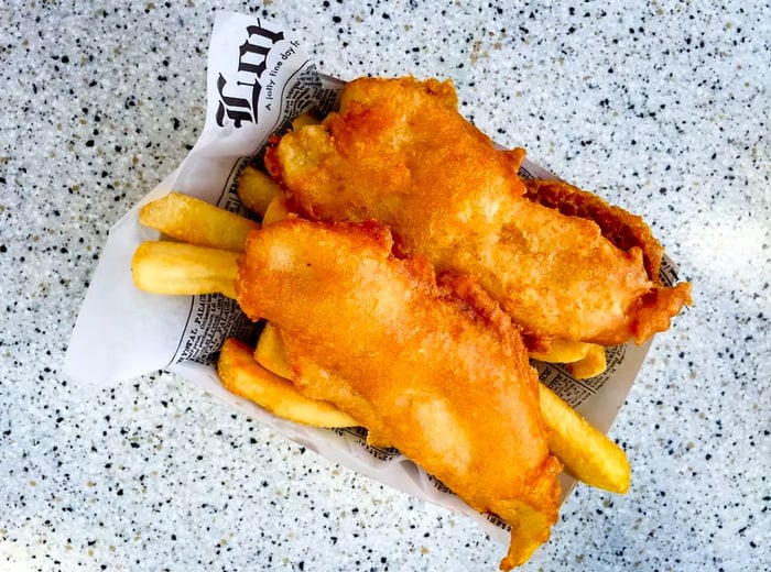 A bird's-eye view of two pieces of fried fish on a bed of fries in a newspaper-lined basket.