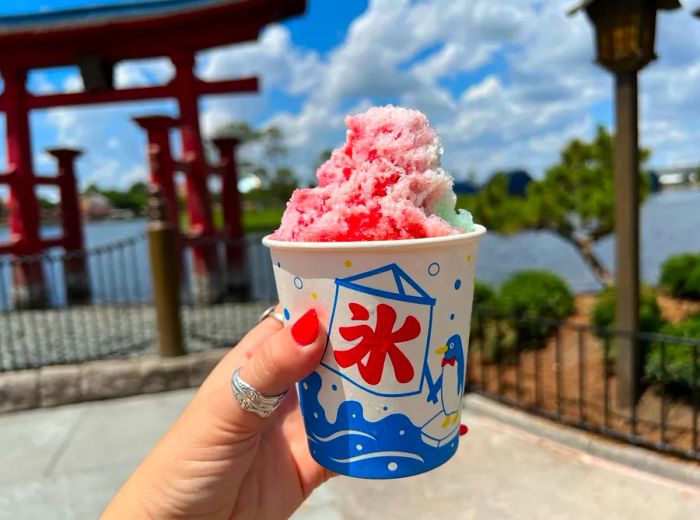 A hand with vibrant nail polish lifts a decorative paper cup filled with shave ice in shades of pink and green, set against a beautifully crafted Japanese gateway arch in the background.