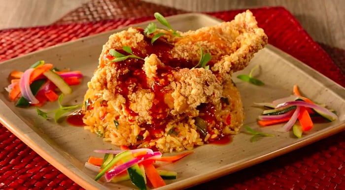 A generous piece of fried chicken topped with sauce and sliced vegetables, served on a plastic plate on a wooden table.