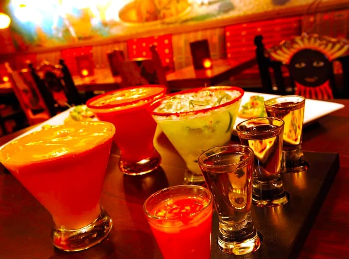 A selection of flavored margaritas and tequila shots set on a table in a dimly lit space with white brick walls.