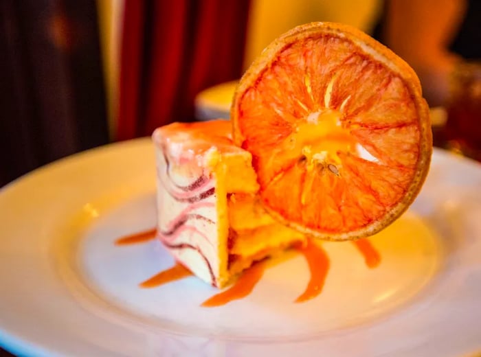 A slice of cake resting in a pool of swirled sauce, topped with a large dried orange slice.