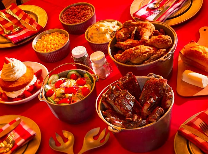 Buckets filled with fried chicken, accompanied by salad, dessert, and sides like mac and cheese and beans, set on a red tablecloth near dinner plates.