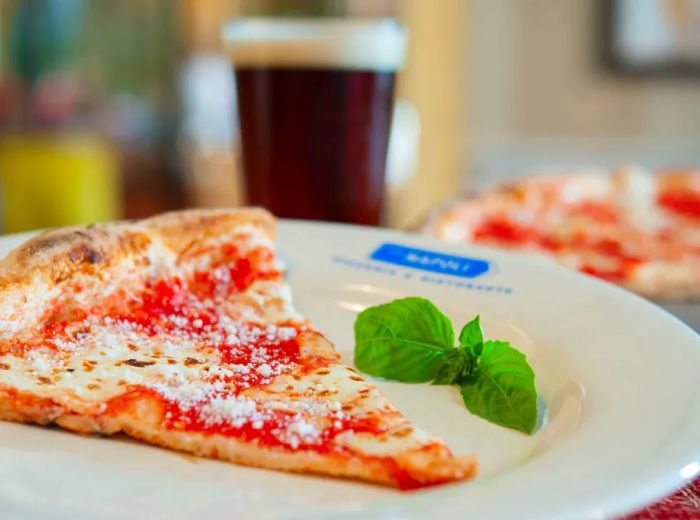 A slice of pizza garnished with a small sprig of basil, with the rest of the pie displayed in the background.