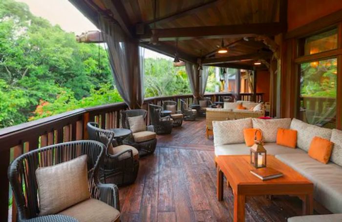 A second-story wooden balcony furnished with patio chairs and couches, surrounded by tall trees for a tranquil atmosphere.