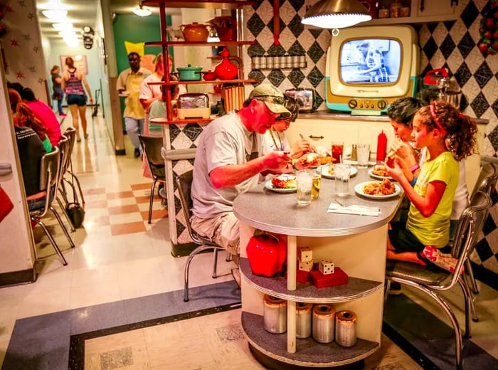 Guests seated in a dining room reminiscent of the 1950s, featuring vintage TV sets, patterned walls, and classic kitchen decor.