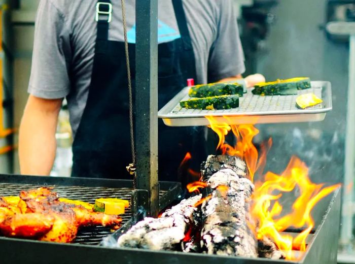 A chef stands over an open wood fire, holding a tray of vegetables while a spatchcocked chicken roasts beside it.