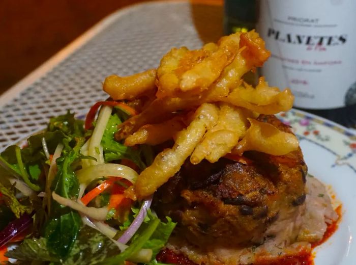 A decorative plate showcases a mound of pastelón crowned with a heap of French fries, accompanied by a salad and a bottle of wine nearby.