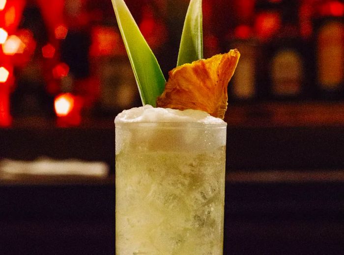 A tall cocktail resting on the bar, adorned with two pineapple fronds and a wedge perched atop. The drink is a milky yellow with a frothy layer. Neon lights illuminate the bar.