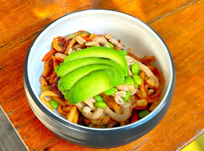 A bowl of noodles garnished with sliced avocados, peanuts, and sauce.