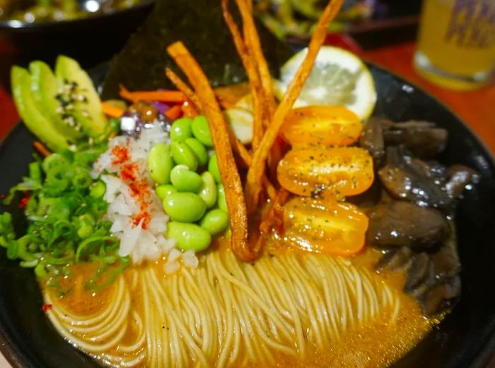 A bowl of ramen featuring curled noodles on one side, adorned with toppings like edamame, scallions, onions, tomatoes, mushrooms, and crunchy sticks.