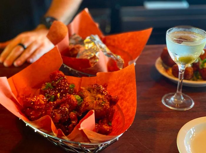 A basket lined with paper holding chicken wings, another basket behind it, and a martini resting on a wooden bar, with a server nearby.