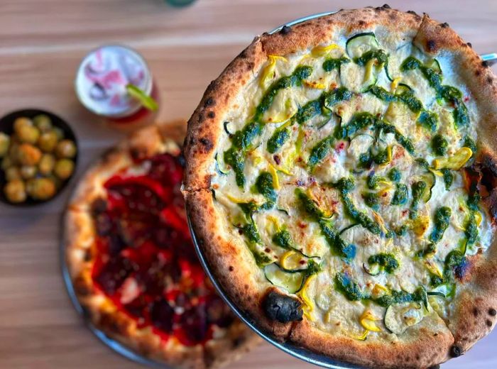 A close-up of one pizza drizzled with green sauce, with another pizza featuring red sauce in the background.