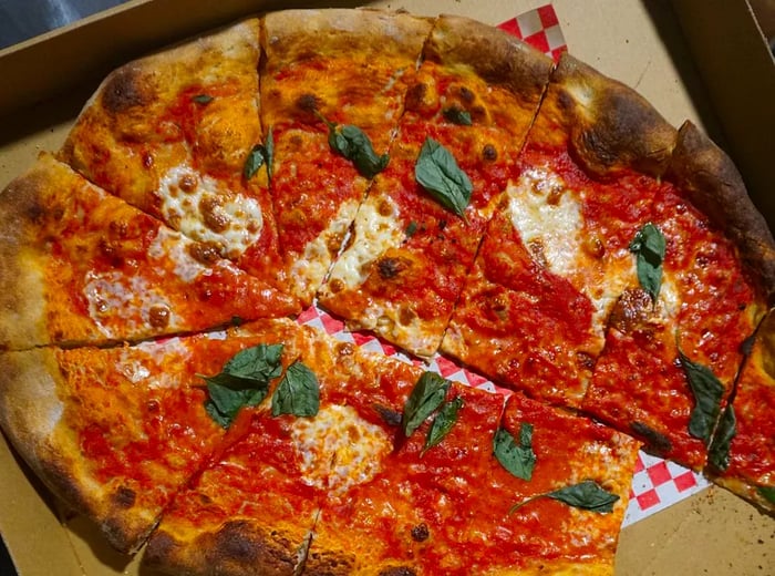 An oval pizza cut into wedges, topped with tomato sauce, generous cheese, and fresh basil leaves, presented in a cardboard box with one slice already taken.