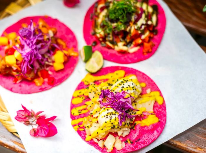 Three tacos featuring a variety of colorful fillings, all served on vibrant pink tortillas.