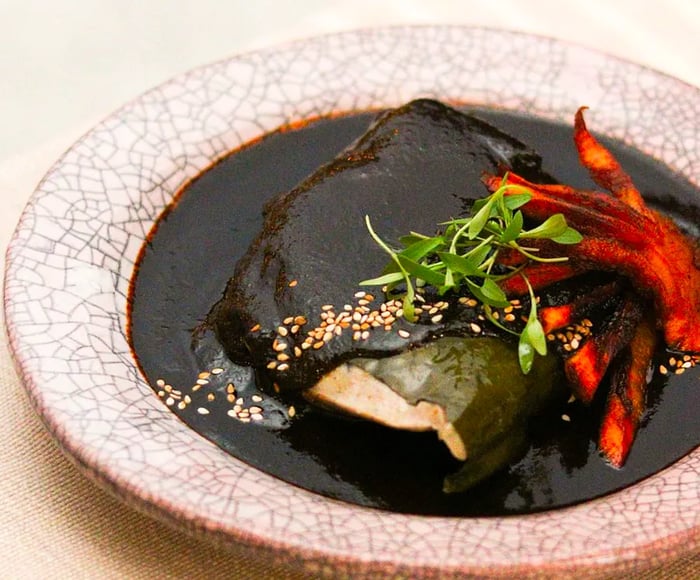 A tamal drenched in dark mole, garnished with sesame seeds and herbs.