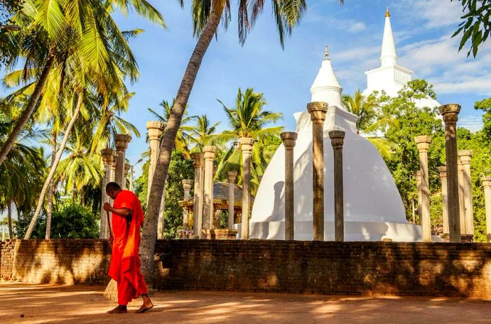 An orange-robed monk sweeps the ground at a temple in Mihintale, Sri Lanka.