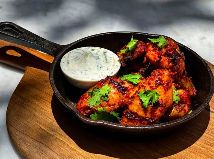 A cast iron skillet brimming with spiced chicken pieces accompanied by a dipping sauce.