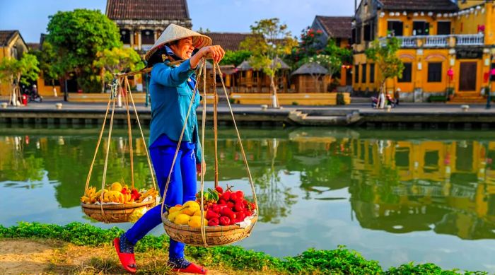 A woman walks along a canal in Hanoi's Old Town, balancing baskets filled with fruits.